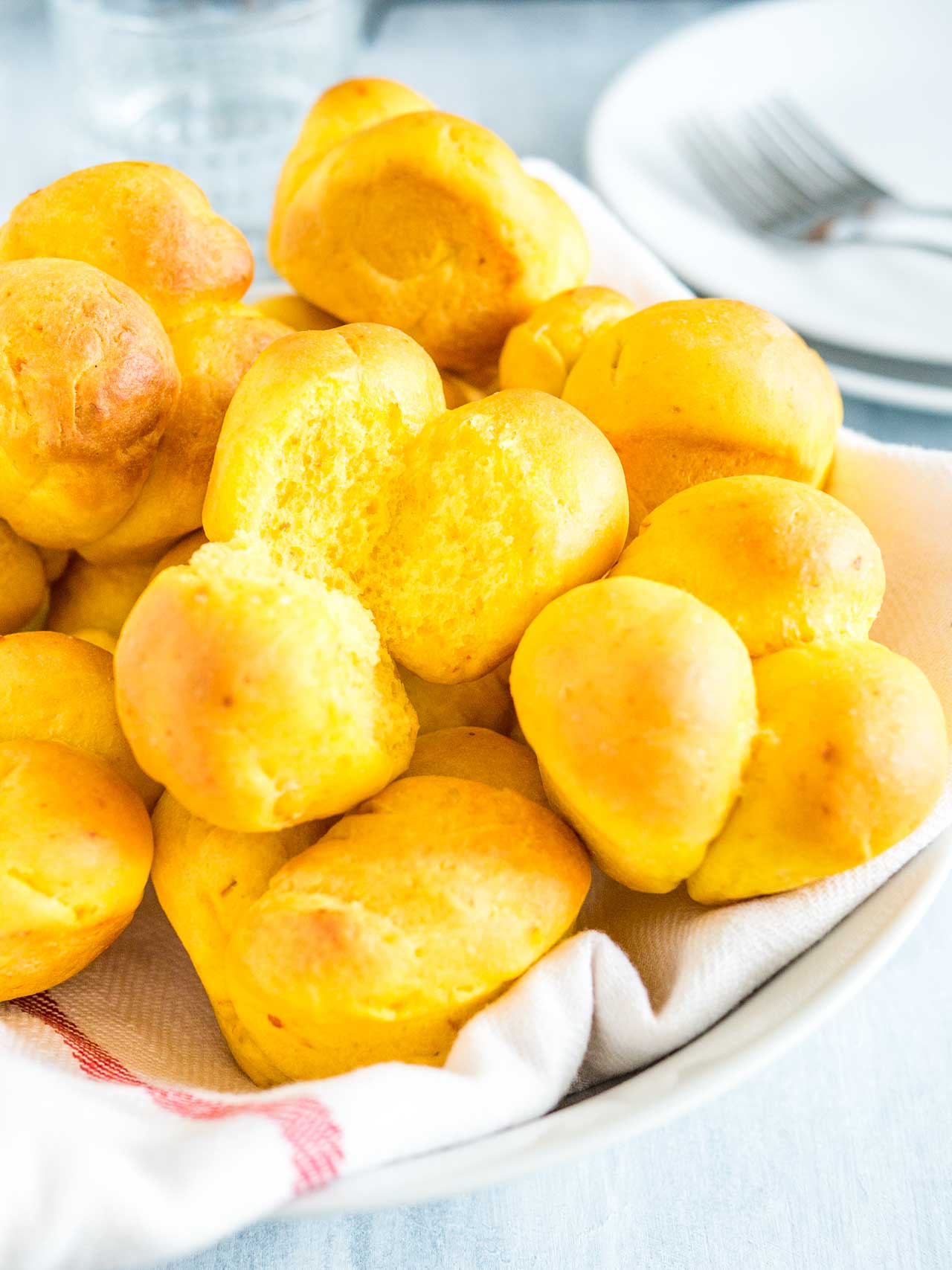 Pumpkin dinner rolls on a white dishtowel with a red stripe on a white plate. One of the rolls has a piece missing and there\'s a stack of white plates with forks in the background.