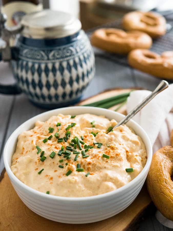 A white bowl of Obatzda (German Beer Cheese Dip) with a spoon, garnished with chives on a wooden cutting board. There\'s a pretzel on a white dishtowel with a red stripe next to it and a beer stein in the background with more pretzels.