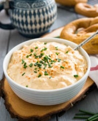 A white bowl of Obatzda (German Beer Cheese Dip) with a spoon, garnished with chives on a wooden cutting board. There's a pretzel on a white dishtowel with a red stripe next to it and a beer stein in the background.
