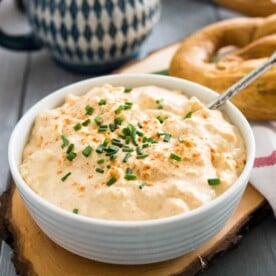 A white bowl of Obatzda (German Beer Cheese Dip) with a spoon, garnished with chives on a wooden cutting board. There's a pretzel on a white dishtowel with a red stripe next to it and a beer stein in the background.
