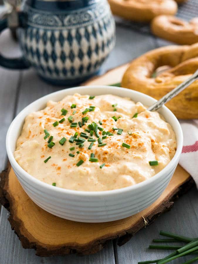 A white bowl of Obatzda (German Beer Cheese Dip) with a spoon, garnished with chives on a wooden cutting board. There\'s a pretzel on a white dishtowel with a red stripe next to it and a beer stein in the background.