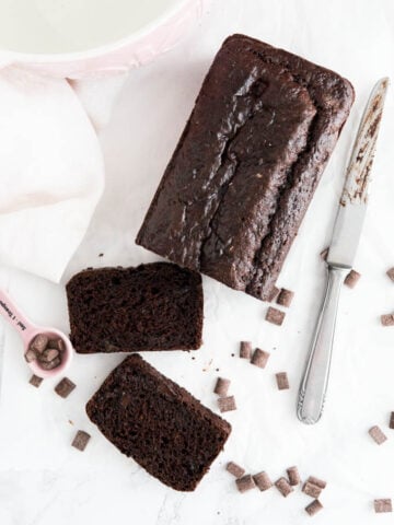 Top-down shot of a double chocolate chip bread with two slices cut off and lying next to it on a marble surface. There are chocolate chips, a pink measuring spoon with chocolate chips, a knife and a white dishtowel next to it.