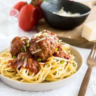 A grey plate with spaghetti and Italian Meatballs with tomato sauce on a white dishtowel. There's a bronze fork next to it and a glass of water, some tomatoes on the vine and a wooden cutting board with a black bowl of grate Parmiggiano in the background.