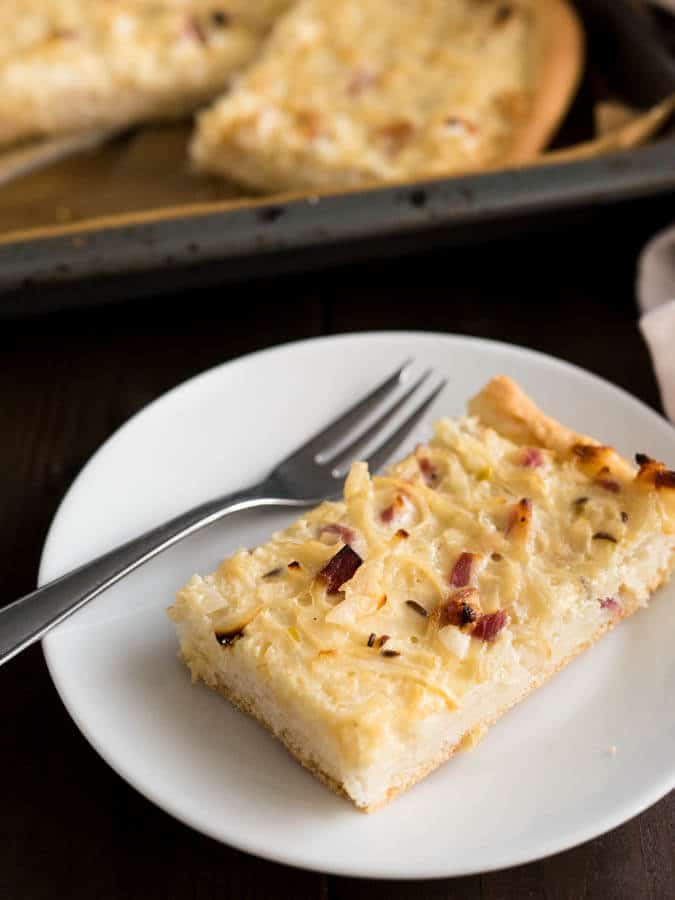 A white plate with a piece of onion tart and a fork. There\'s a baking sheet with more of the tart in the background.
