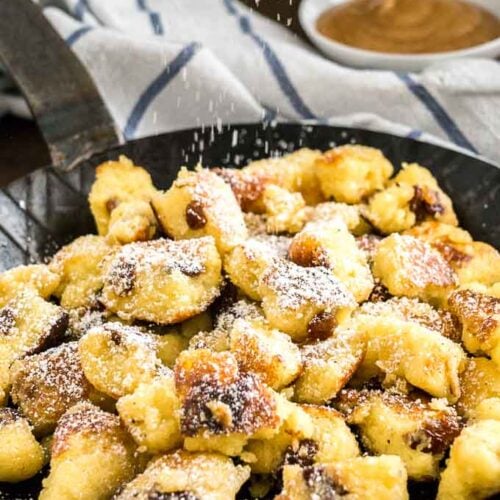 A pan with Kaiserschmarrn with powdered sugar on top on a white and blue dish towel with a white bowl of apple sauce in the background
