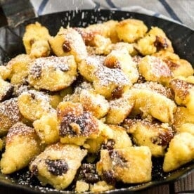 Close-up of a pan with Kaiserschmarrn with powdered sugar on top on a white and blue dish towel with a white bowl of apple sauce in the background