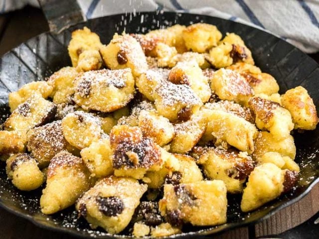 Close-up of a pan with Kaiserschmarrn with powdered sugar on top on a white and blue dish towel with a white bowl of apple sauce in the background