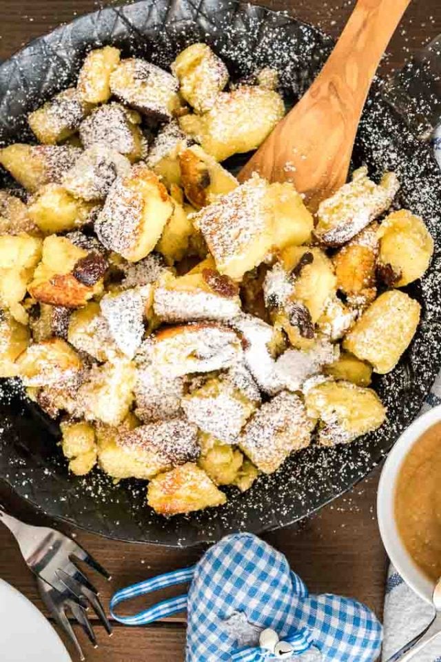 A pan of Kaiserschmarrn with raisins in a pan, topped with powdered sugar, with a wooden spatula in it. There\'s a fork, a white and blue plaid towel and a small white bowl of apple sauce next to it.