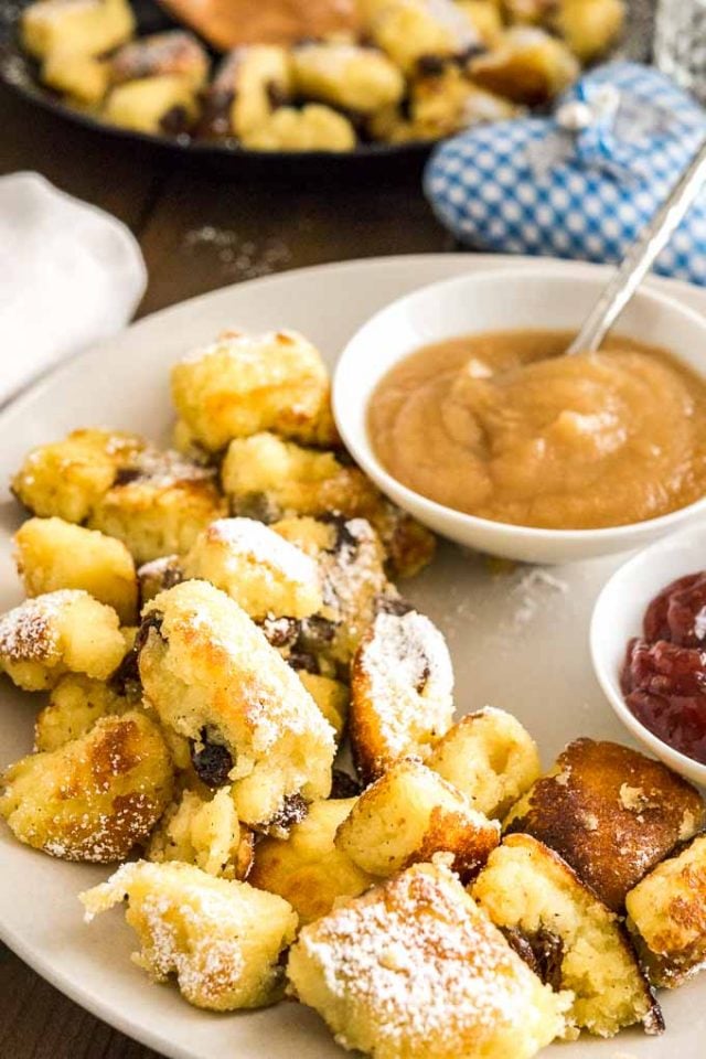 A plate with Kaiserschmarrn topped with powdered sugar with two small white bowls, one with apple sauce, one with plum puree on a wooden table.