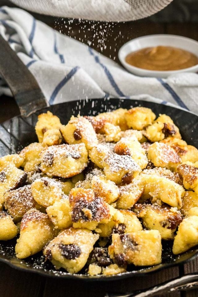 A pan with Kaiserschmarrn with powdered sugar on top on a white and blue dish towel with a white bowl of apple sauce in the background