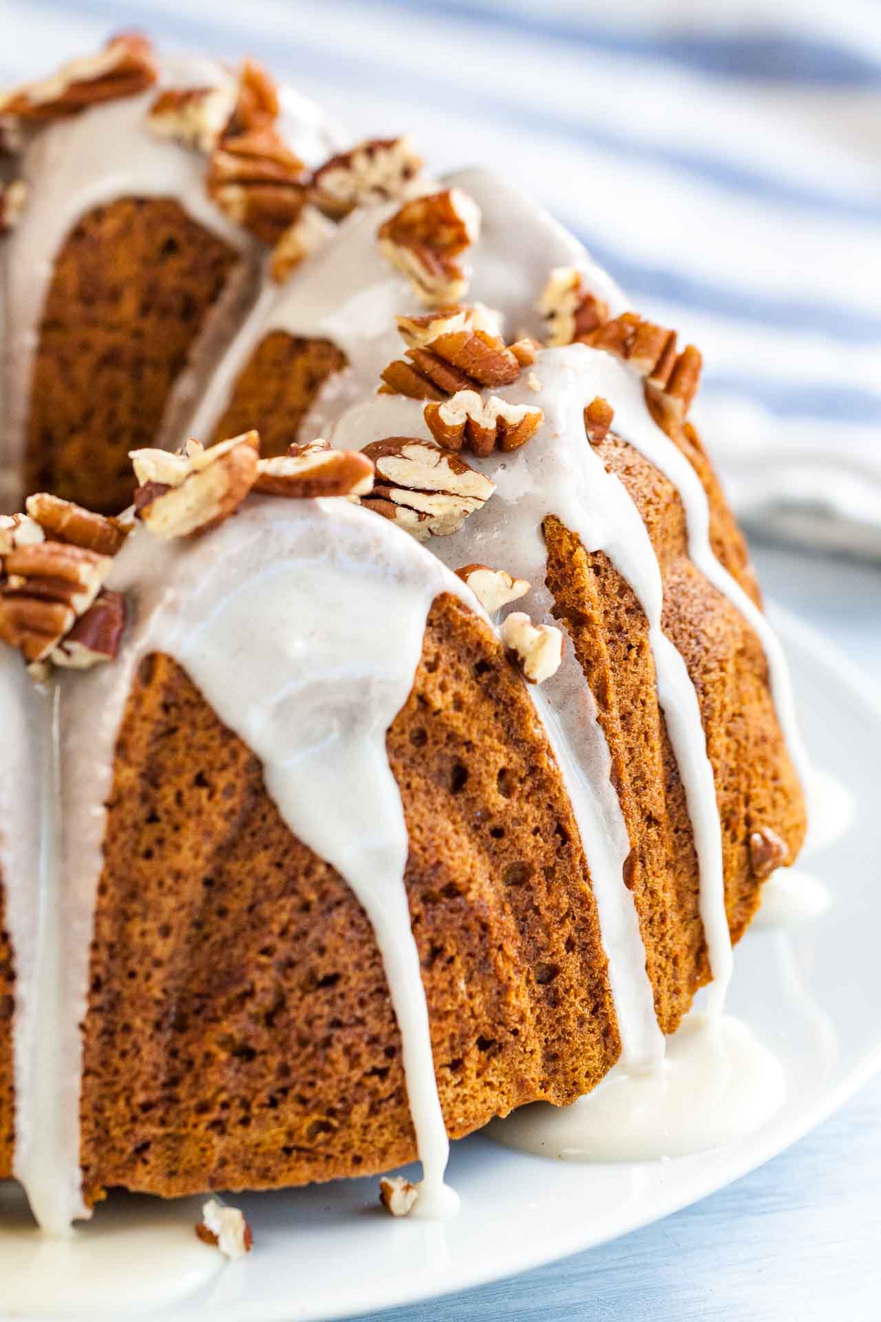 Close-up of a pumpkin bundt cake with maple glaze and toasted pecans on a white plate next to a white and blue dishtowel.