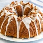 A pumpkin bundt cake with maple glaze and toasted pecans on a white plate next to a white and blue dishtowel.