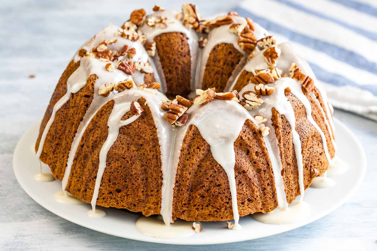 A pumpkin bundt cake with maple glaze and toasted pecans on a white plate next to a white and blue dishtowel.