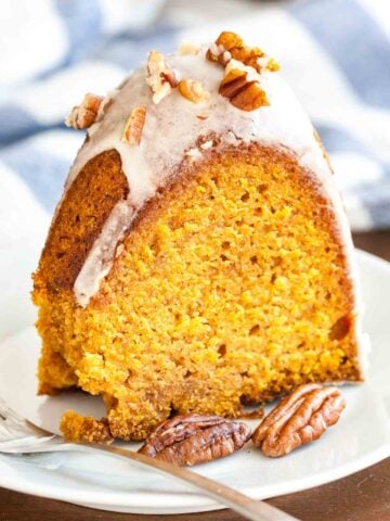 A slice of pumpkin bundt cake with maple glaze and pecans on a small white plate with a fork next to a white and blue dishtowel.