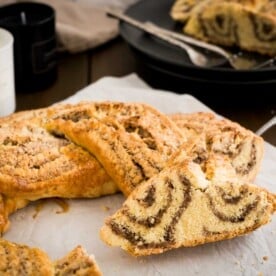 A nut roll, with braided strands of dough with hazelnuts on white parchment paper with a slice cut off and leaning against it. There's a black plate with two forks and more of the nut roll in the background.