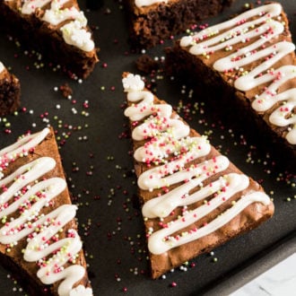 A black baking sheet with gingerbread Christmas brownies with a triangular shape and waves of frosting and sprinkles to make it look like small Christmas trees.