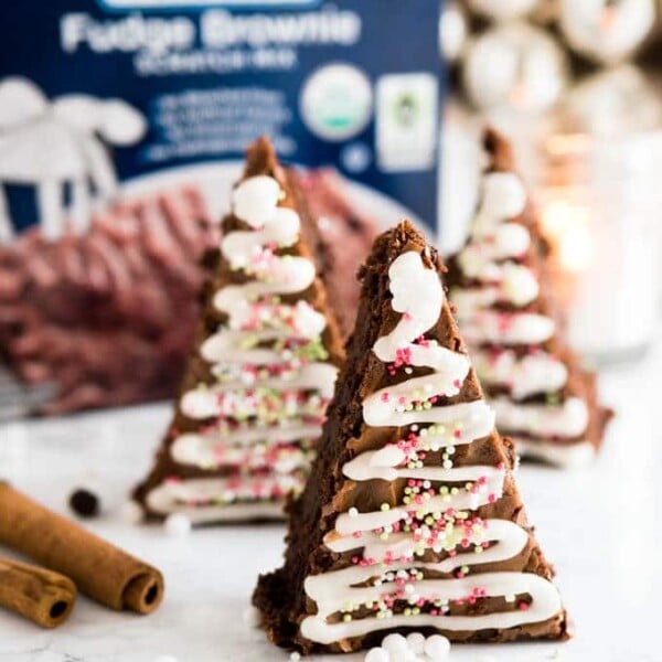 Gingerbread Christmas brownies with a triangular shape and waves of frosting and sprinkles to make them look like small Christmas trees standing in front of a box of fudge brownie mix. There are cinnamon sticks next to it.