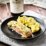 A black plate with parmesan-crusted salmon and mashed potatoes on a grey dishtowel on a wooden table. There's a glass and a bottle of wine next to it.