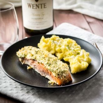 A black plate with parmesan-crusted salmon and mashed potatoes on a grey dishtowel on a wooden table. There's a glass and a bottle of wine next to it.
