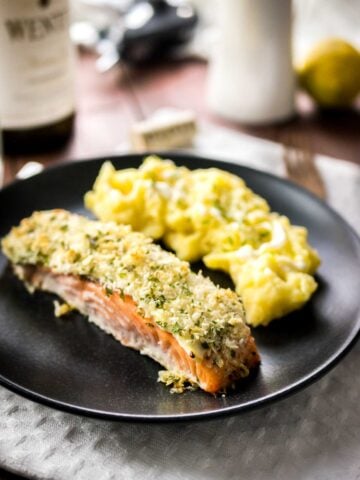 Close-up of a black plate with parmesan-crusted salmon and mashed potatoes on a grey dishtowel on a wooden table. There's a bottle of wine next to it.