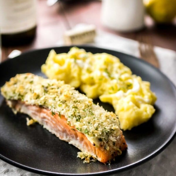 Close-up of a black plate with parmesan-crusted salmon and mashed potatoes on a grey dishtowel on a wooden table. There's a bottle of wine next to it.