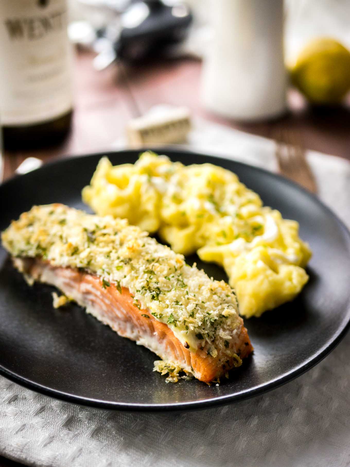 Close-up of a black plate with parmesan-crusted salmon and mashed potatoes on a grey dishtowel on a wooden table. There\'s a bottle of wine next to it.