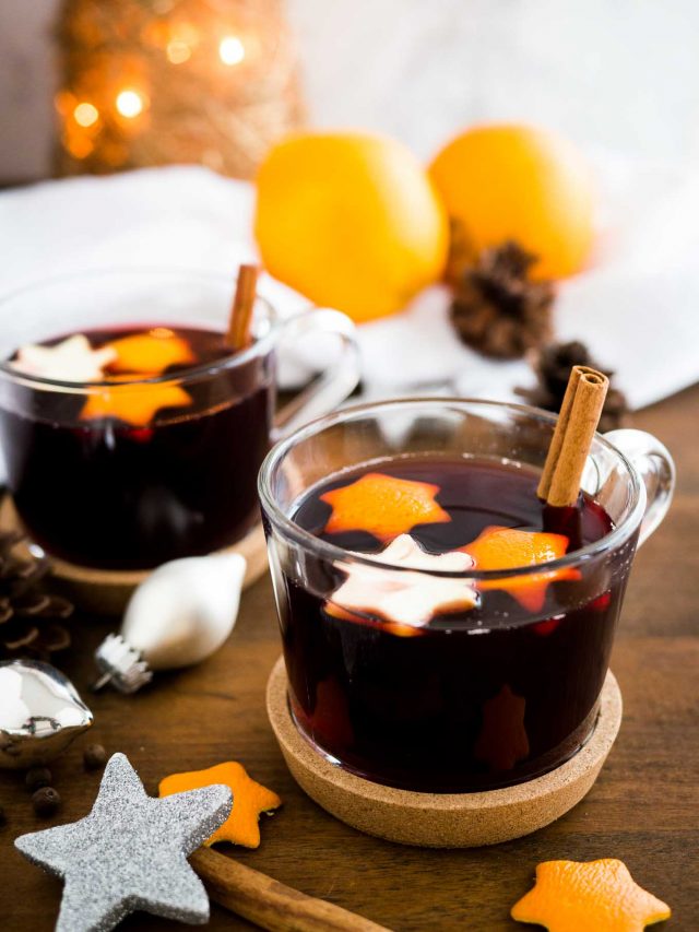 Two glass cups of German Christmas Punch (Kinderpunsch) with stars from orange peel and cinnamon sticks on a wooden table. There are Christmas ornaments next to it and a white dishtowel with oranges and pinecones in the background.