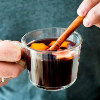 Two hands holding up a glass cup of German Christmas Punch (Kinderpunsch) with stars from orange peel. One hand is holding a cinnamon stick in it.