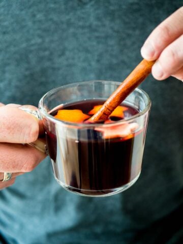 Two hands holding up a glass cup of German Christmas Punch (Kinderpunsch) with stars from orange peel. One hand is holding a cinnamon stick in it.