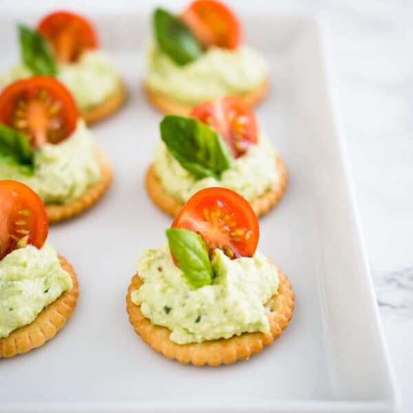 Feta tomato bites on a white plate on a marble surface. The bites consist of a cracker, with feta-basil spread topped with a tomato and basil.