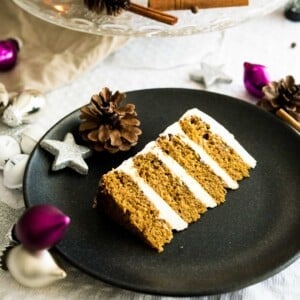 A black plate with a slice of gingerbread cake on a grey tableclother. There are pinecones, silver and pink Christmas ornaments and a glass platter with cinnamon sticks next to it.