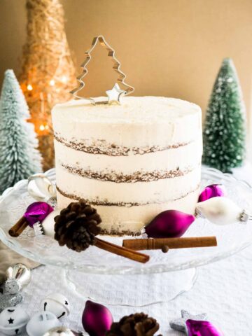 A glass cake platter with gingerbread cake, with cinnamon sticks, pinecones and Christmas ornaments. There are two little pine trees and a gold Christmas tree ornament in the background.