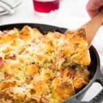 A cast-iron pan with ham and cheese breakfast casserole. A hand is digging into it with a spatula. In the background, there's a stack of white plates with cutlery, a white dishtowel and a glass of fruity tea.
