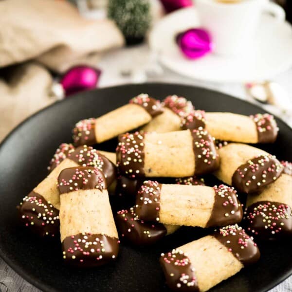 Close-up of A black plate of maple hazelnut cookies, with both ends dipped in chocolate and sprinkles on a grey tablecloth. There are silver and pink Christmas ornaments lying next to it and a white cup with espresso in the background.