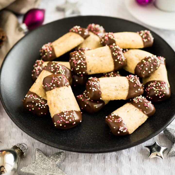 A black plate of maple hazelnut cookies, with both ends dipped in chocolate and sprinkles on a grey tablecloth. There are silver and pink Christmas ornaments lying next to it.