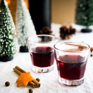 Two glasses of mulled wine liqueur on a grey tablecloth with cinnamon sticks. There are pinecones and little pine trees in the background.