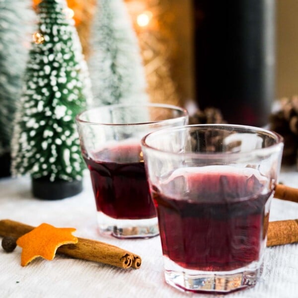Close-up of two glasses of mulled wine liqueur on a grey tablecloth with cinnamon sticks. There are pinecones and little pine trees in the background.
