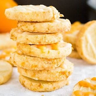 A stack of orange almond cookies on a white tablecloth with more of the cookies next to it and an orange in the background.