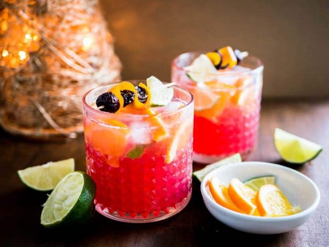 Two glasses of orange cranberry Moscow mule with fruit skewers. There\'s a small white bowl of lime and orange wedges as well as halved limes next to it and a Christmas ornament in the background.