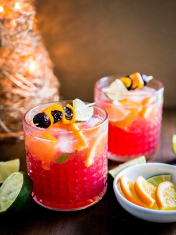 Two glasses of orange cranberry Moscow mule with fruit skewers. There's a small white bowl of lime and orange wedges as well as halved limes next to it and a Christmas ornament in the background.