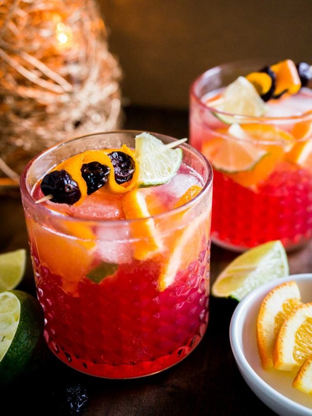 Close-up of two glasses of orange cranberry Moscow mule with fruit skewers. There\'s a small white bowl of lime and orange wedges as well as halved limes next to it and a Christmas ornament in the background.