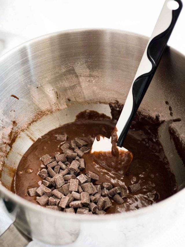 A stainless steel mixing bowl with chocolate batter and chocolate chips, with a black and white spatula in it.