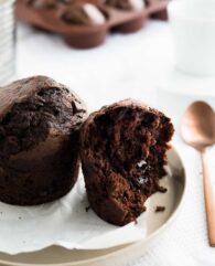 A chocolate banana muffin next to half a chocolate muffin with a molten chocolate core on a grey plate with parchment paper next to a bronze spoon with a glass and more muffins in the background.