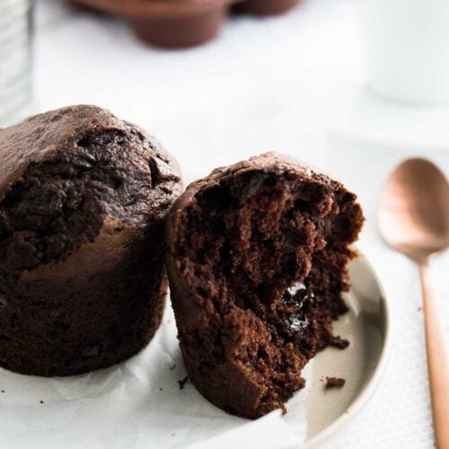 A chocolate banana muffin next to half a chocolate muffin with a molten chocolate core on a grey plate with parchment paper next to a bronze spoon with a glass and more muffins in the background.