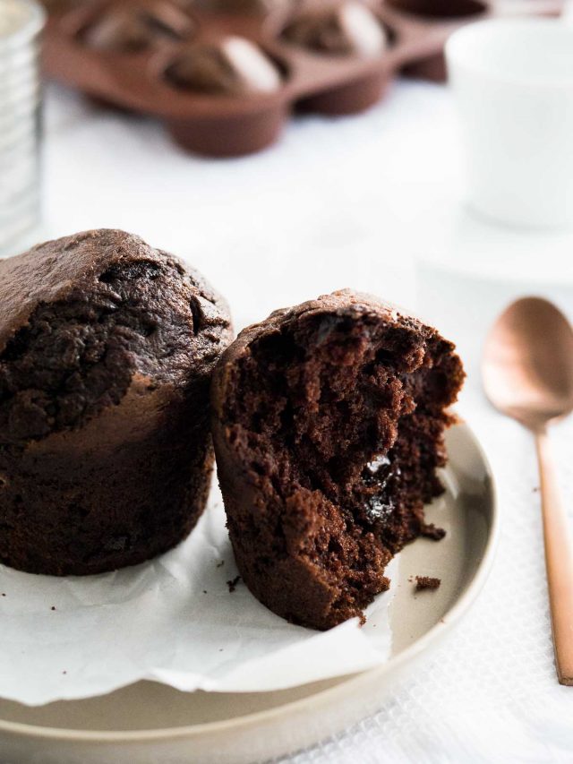 A chocolate banana muffin next to half a chocolate muffin with a molten chocolate core on a grey plate with parchment paper next to a bronze spoon with a glass and more muffins in the background.