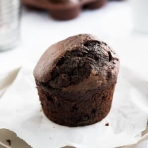 A chocolate banana muffin on a grey plate with parchment paper next to a bronze spoon with a glass and more muffins in the background.
