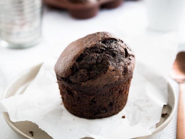 A chocolate banana muffin on a grey plate with parchment paper next to a bronze spoon with a glass and more muffins in the background.