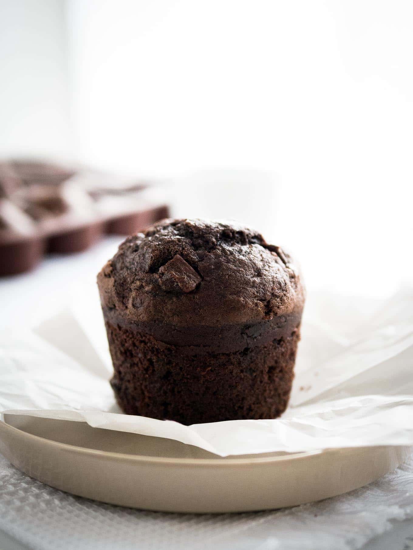 Close-up of a chocolate banana muffin on a grey plate with parchment paper with more muffins in the background.