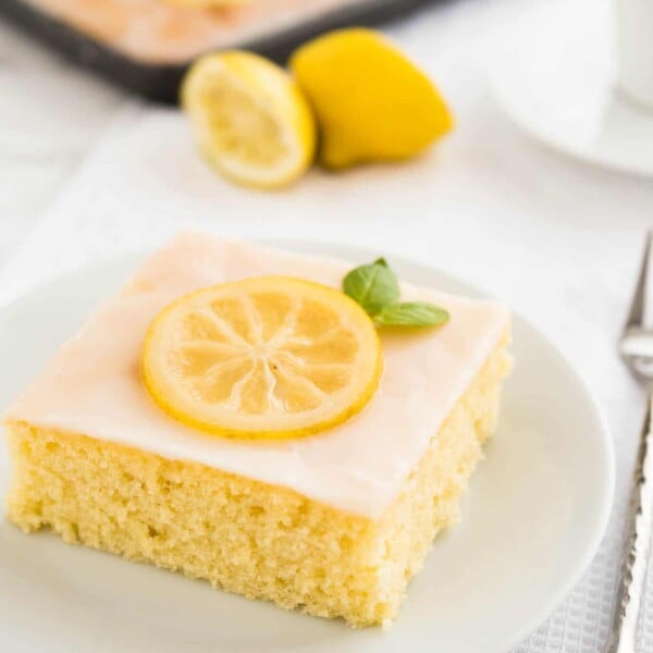 A slice of lemon sheet cake with lemon frosting, a slice of lemon and mint on a white plate on a white dishtowel. There are two lemon halves a white teacup and the rest of the sheet cake in the background and a fork next to it.