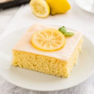 A slice of lemon sheet cake with lemon frosting, a slice of lemon and mint on a white plate on a white dishtowel. There are two lemon halves a white teacup and the rest of the sheet cake in the background and a fork next to it.